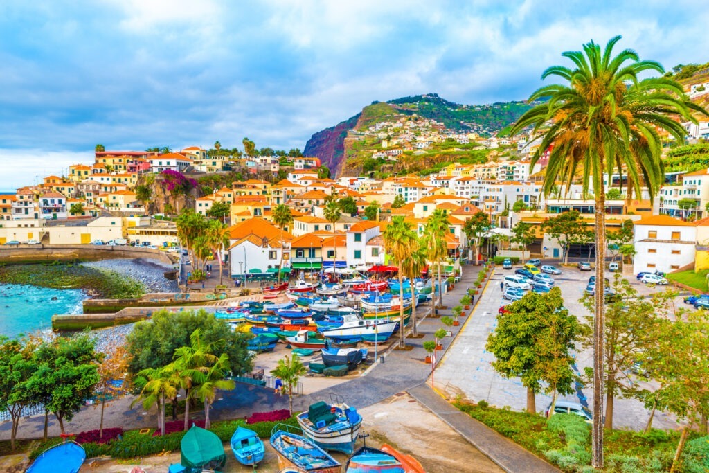 Blick auf das bunte Küstendorf Camara de Lobos auf Madeira mit Palmen, Fischerbooten und farbenfrohen Häusern vor grüner Hügellandschaft.