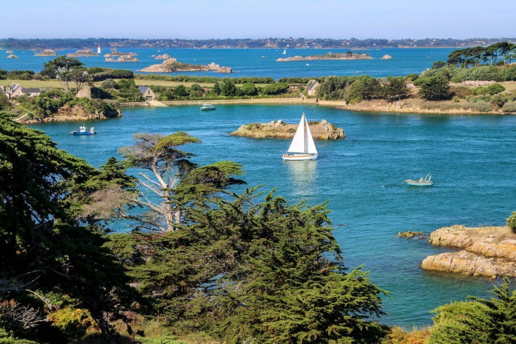 Malerische Küstenlandschaft in Frankreich mit türkisfarbenem Wasser, Segelboot, Felseninseln und Pinien – ideale Atmosphäre für FKK-Strände.