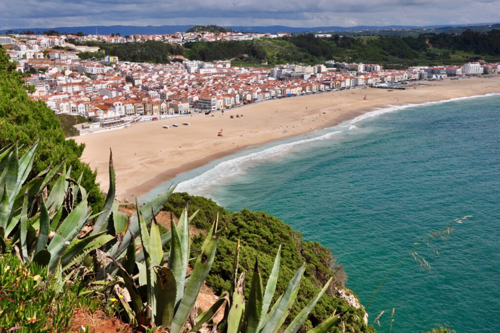 Blick über Praia da Nazaré mit Promenade, Strand, Meer und dahinterliegender Stadt in der Abendsonne