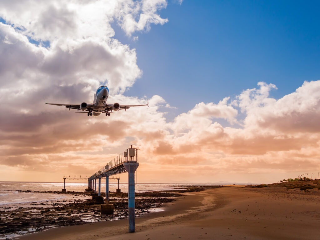 Airplane approaching beach on Lanzarote island, Spain. Landing plane over ocean against sunset sky seen from view point Mirador de Acercamiento, Canary islands WeitlĂ€ufiger, ruhiger Sandstrand mit tĂŒrkisblauem Meer und beeindruckender SteilkĂŒste auf Gran Canaria â ideal fĂŒr FKK-Urlaub.