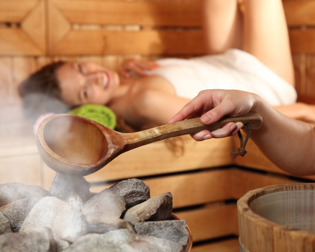 Zwei lächelnde Frauen liegen entspannt auf Handtüchern in einer Holzsauna im Warmbad Villach.