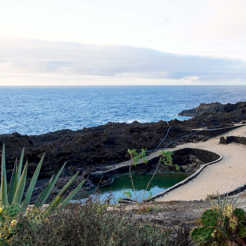 Calm natural pool nestled among dark volcanic rock and sandy sunbathing areas in a naturist village by the ocean, offering relaxed, carefree seaside wellness and swim escape FKK-Strand Charco del Palo auf Lanzarote – natürliche Meeresbecken zwischen Lavafelsen, beliebtes Ziel für FKK-Urlauber auf den Kanaren.