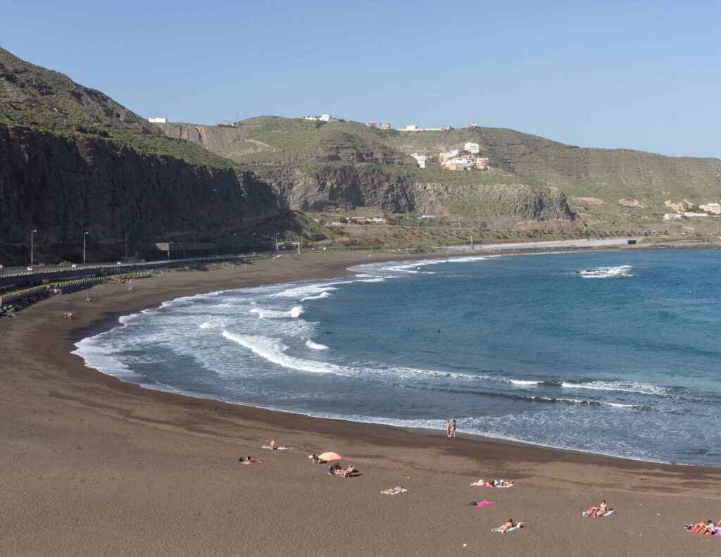 Playa de arena negra con cielo azul y montañas Weitläufiger Sandstrand von Playa del Inglés auf Gran Canaria mit Meerblick, Weitläufiger Sandstrand von Playa del Inglés auf Gran Canaria mit Meerblick, Sonnenschirmen und FKK-Bereich nahe den Dünen von Maspalomas.